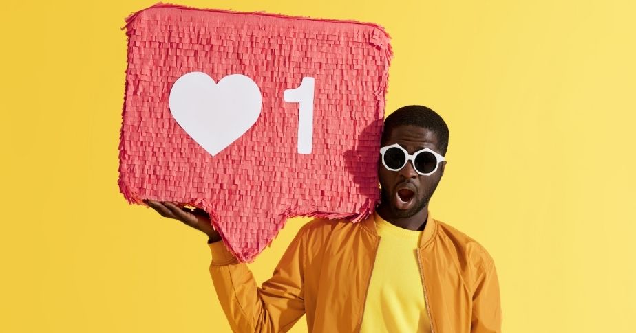 Com um plano de fundo amarelo, um homem negro vestindo uma jaqueta laranja segura um imenso ícone, que representa uma curtida no feed do Instagram ou do Insta Story.