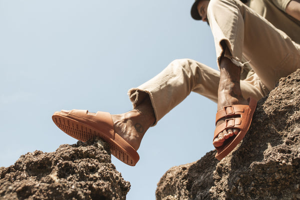 Foto de artigo sobre Linus mostra uma pessoa sentada em pedras, apoiando os pés na pedra também. A pessoa veste uma calça cáqui e um par de sandálias Linus de cor marrom. A foto foi tirada de um ângulo inferior ao modelo para destacar as sandálias.