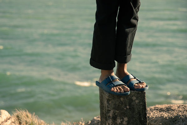 Foto de artigo sobre a loja Linus mostra uma pessoa em uma praia, de pé sobre um toco de madeira. A foto foca nos pés da pessoa, que está usando uma calça comprida e uma sandália Linus azul claro. Ao fundo, há o mar azul.