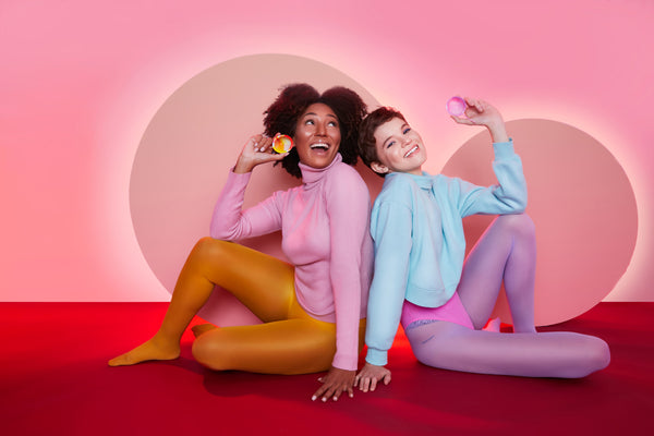 Foto do empreendedorismo feminino mostra duas mulheres sentadas no chão, uma de costas para a outra, ambas segurando um disco menstrual. As duas mulheres estão sorrindo. No fundo, há dois círculos em alusão à Lua.