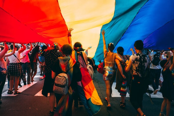 Foto de artigo sobre o mês LGBTQIA+ mostra diversas pessoas em uma avenida coberta pela bandeira do orgulho LGBTQIA+.