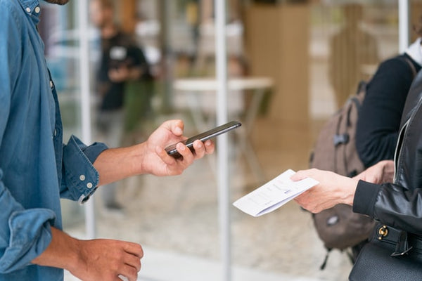 Foto mostra uma pessoa à direita segurando um papel, enquanto à esquerda outra pessoa usa o celular para escanear o que está no papel (meios de pagamento online).