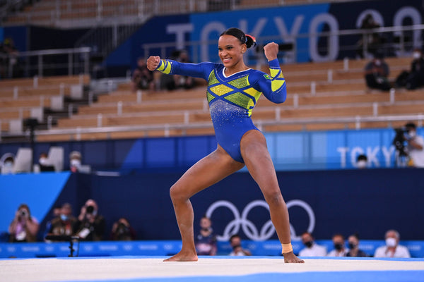 Ao centro, a ginasta Rebeca Andrade está de pé, com os braços erguidos, durante apresentação de solo nos Jogos Olímpicos de Tóquio. Ao fundo, arquibancadas com poucas pessoas e o símbolo dos Jogos Olímpicos.