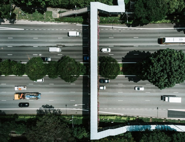 Foto em alusão ao tráfego pago mostra uma avenida de mão dupla, vista de cima. Na avenida, há diversos carros e caminhões. Entre as duas pistas da avenida, há árvores verdes. Sobre a pista, há uma passarela para pedestres.