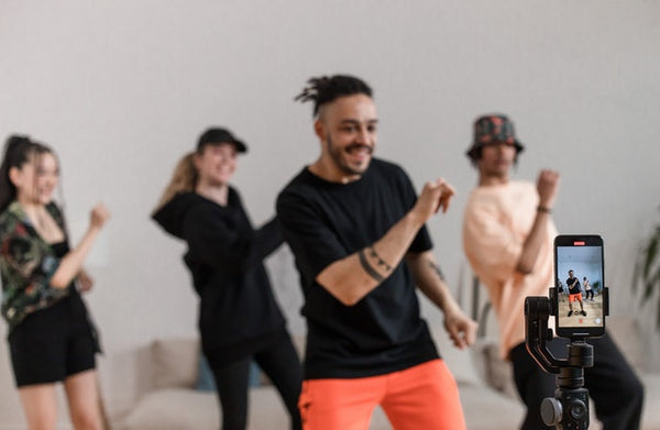 Foto mostra quatro pessoas, vestindo roupas casuais, dançando de frente para um celular em um tripé. O celular está com a câmera ligada e mostra a imagem que está sendo filmada para o TikTok Business