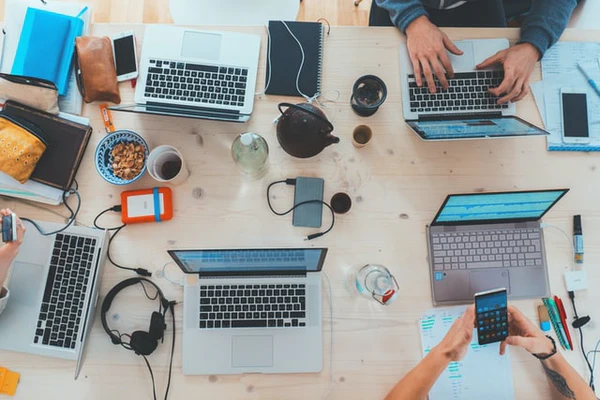 Imagem mostra pessoas sentadas à mesa, todas diante de laptops e smartphones.