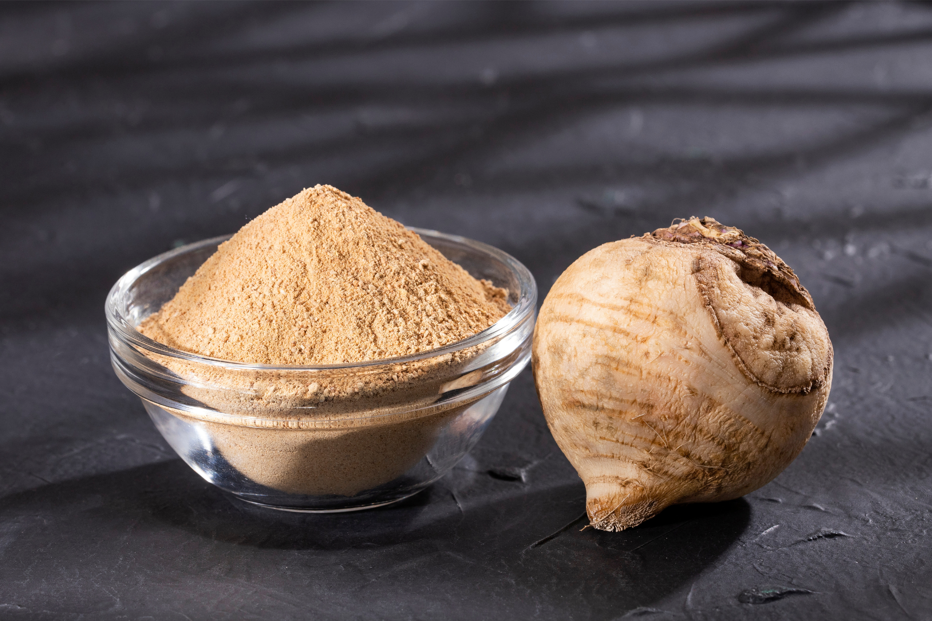 Glass bowl filled with maca root powder beside a whole maca root on a dark, textured background.