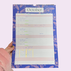 A full page of the Holchester Designs Household Planner is open - at October - and the columns are completed for the family to use. It is being held on the left by a hand in a pink sleeve, and the background is a pale lilac.