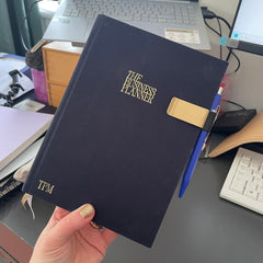 A left hand is holding up a navy-blue linen bound planner book.  In gold on the front is written The Business Planner, and it is a planner from The Productivity Method.