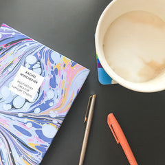 A blue marbled notebook with a personalised panel - Rachel Winchester, Holchester Creative Support Studio - is on a grey desk next to a cup of coffee.