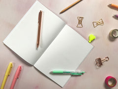 an open A5 notebook lies flat on a table, with pens, highlighters and paperclips scattered around it.