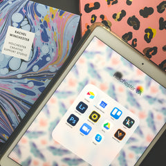 An ipad screen is on, showing a menu with a nunber of creative apps used for running a business.  Next to it is a marble notebook with Rachel Winchester, Holchester Creative Support Studio written on the front.