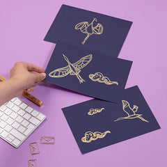Three foiled prints featuring the gold foil migrating cranes - each different in either a standing or flying pose.  A hand is coming from the left to hold up the middle print, with all three leaning on  a lilac desk.