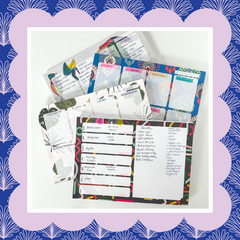 4 planner pads are piled on each other, at angles, on a white desk. There is a lilac scallop edge detail around the photograph and blue pattern behind.