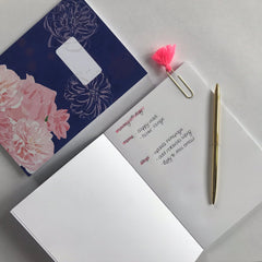 An open, blank paged notebook is lying on a white table with a pen across it.  Next to it at a 90 degree angle to it is a closed floral notebook.