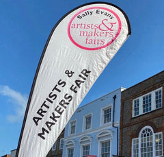 A sail-type banner is outside infront of old buildings and a clear blue sky.  It reads Artists and Makers Fair, with the Sally Evans logo at the top.