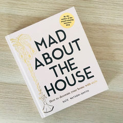 the book Mad about the House is lying on a wooden tabletop so you can see the front cover with the large title and a  gold foil plant to its left.