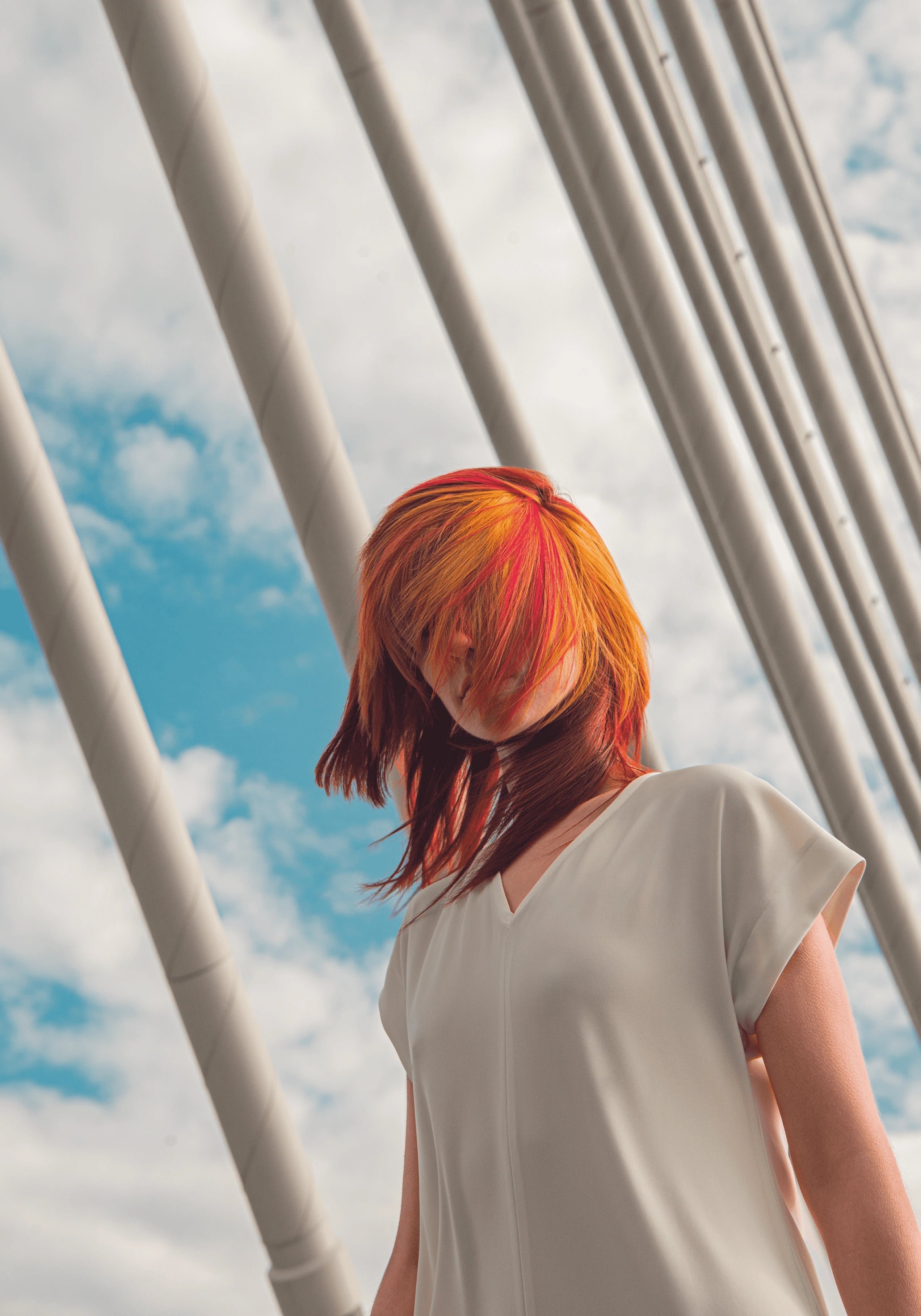 Model mit rot-orangem Haar im weißen Kleid, posiert unter Brückenseilen vor Himmel.