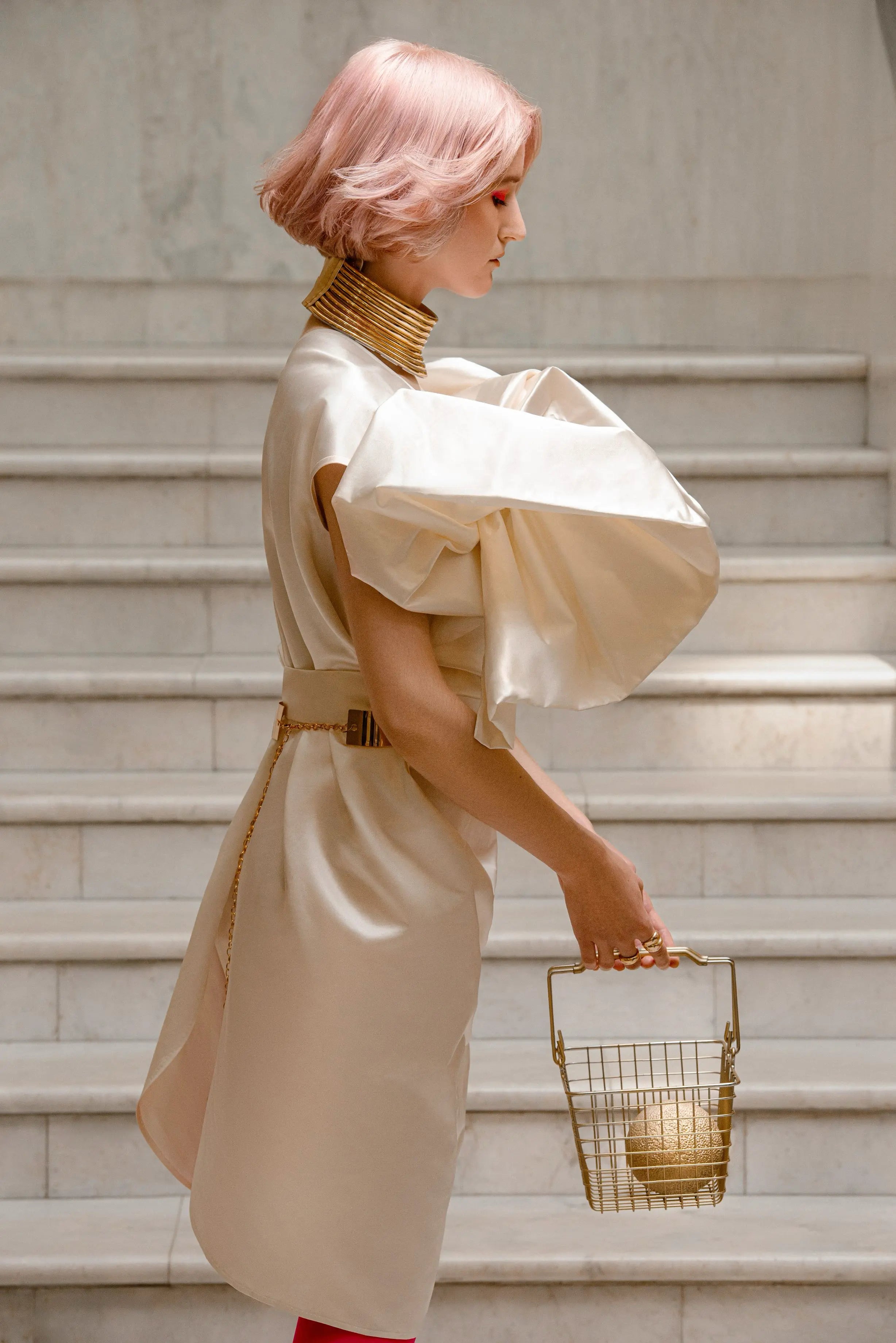 Model mit rosa Bob und goldenem Collier trägt ein weißes Kleid mit großen Puffärmeln und hält einen goldenen Korb.