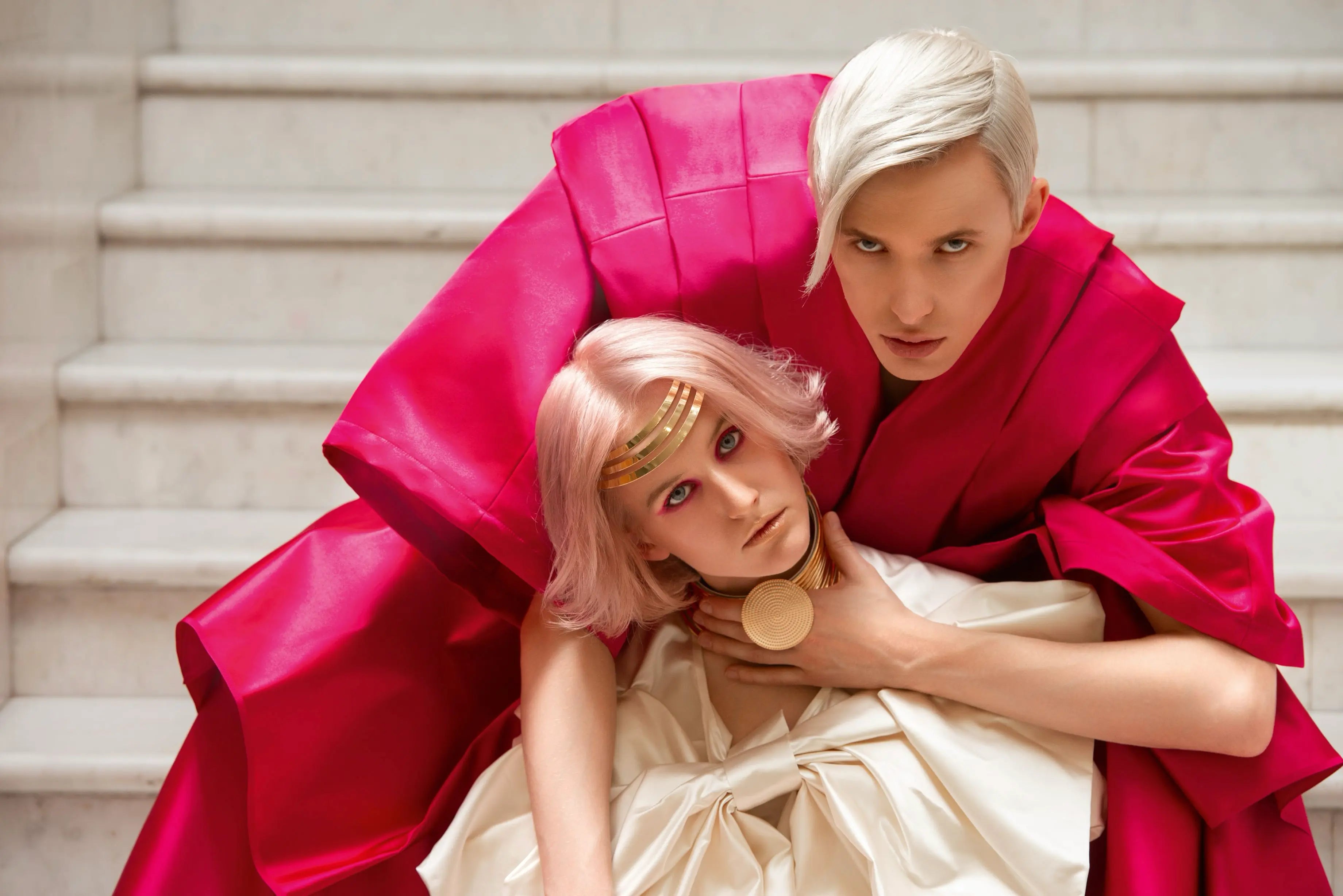 Model im roten Gewand umschließt ein weiteres Model mit rosa Bob und goldenem Accessoire in dramatischer Pose.