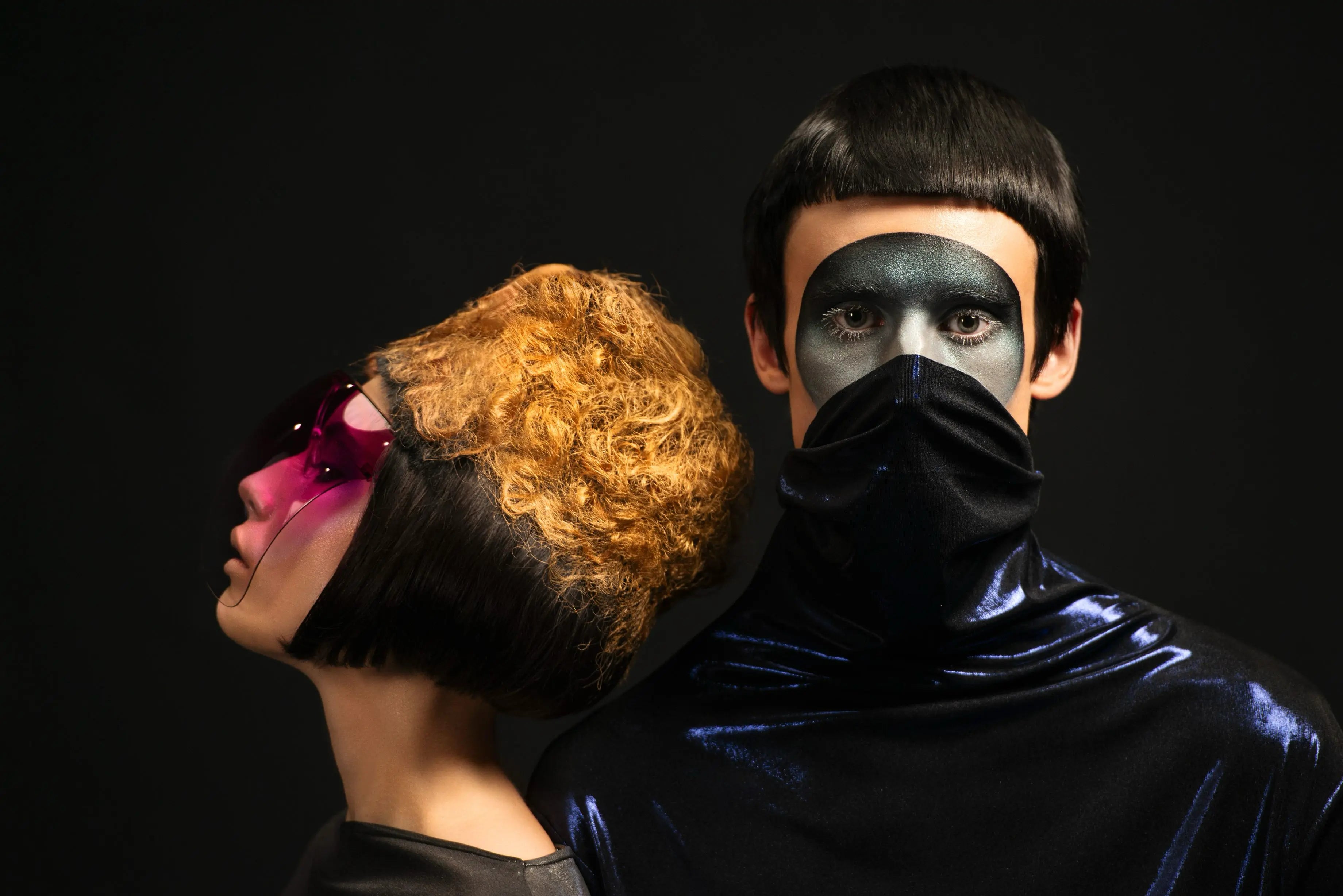 Zwei Personen in schwarzem futuristischem Outfit, eine mit pinker Brille, die andere mit schwarzer Gesichtsmaske.