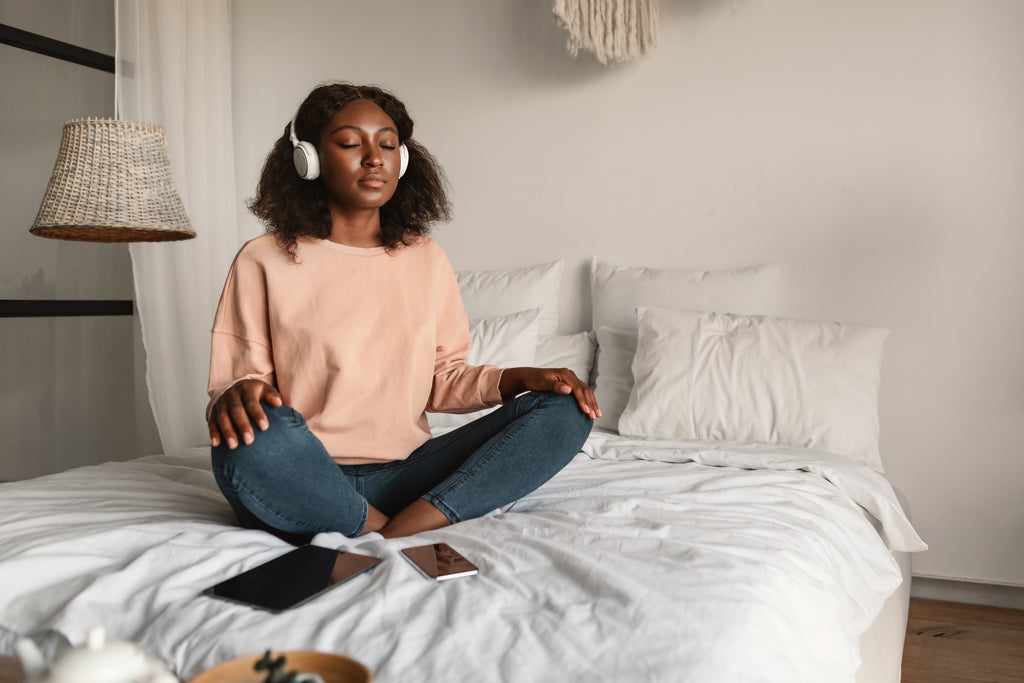 Peaceful Woman Meditating With Eyes Closed Wearing Headphones Sitting In Lotus Position, Listening To Yoga Instructor Via Digital Tablet Relaxing In Bedroom At Home