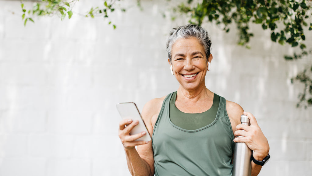 woman outside in her workout clothes holding a water bottle and phone smiling
