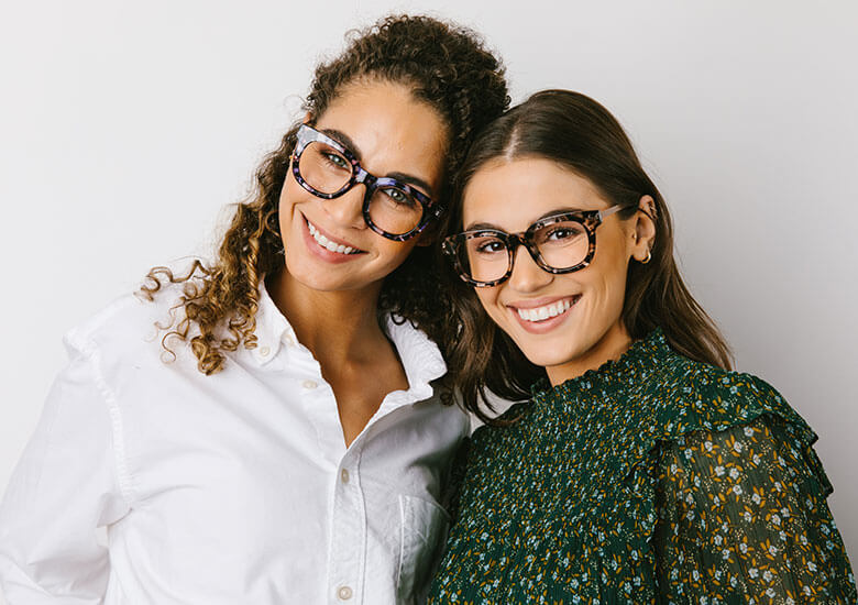 Two woman standing in front of black canvas smiling and looking at camera. Both woman are wearing Peepers Reading Glasses Celeste