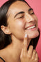 A young woman applies a blemish patch to her chin with a smile