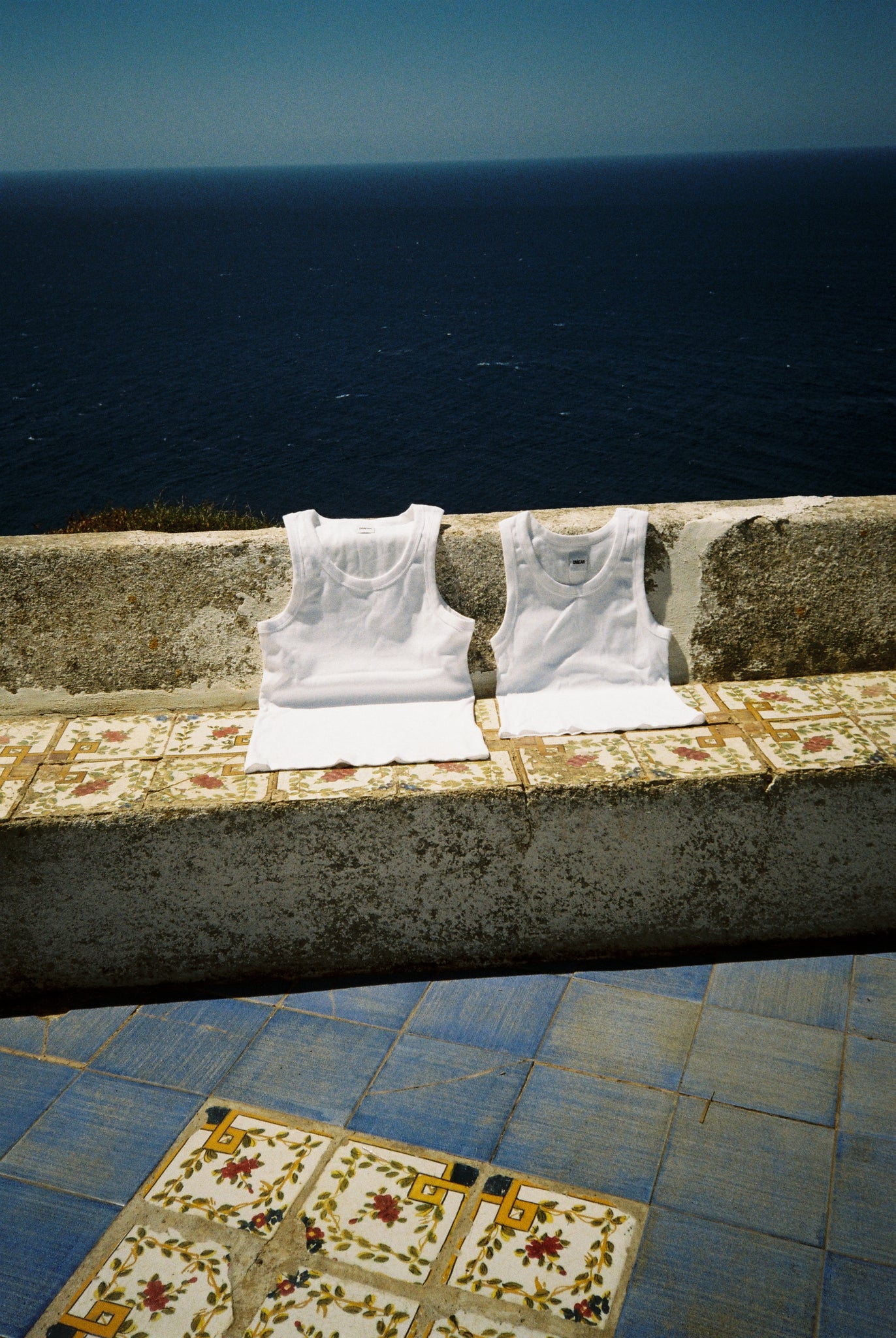Débardeurs en coton blanc OMEAR, séchant au soleil sur un banc en pierre face à la mer à Pantelleria
