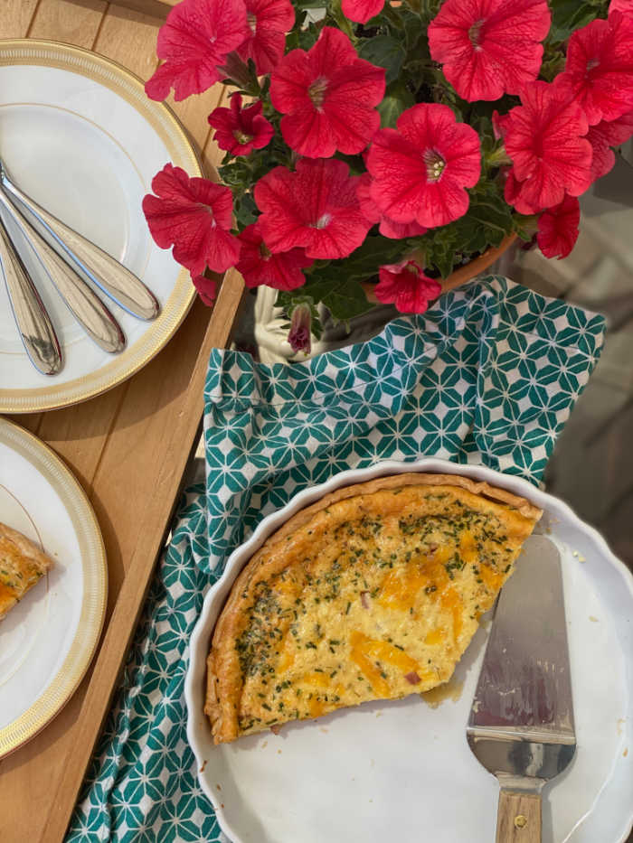 Overhead photo of quiche in pan with spatula, flowers and plates