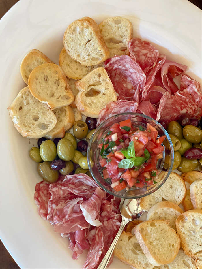 Overhead photo of an antipasto platter with toast, sliced meat, olives, tomato bruschetta