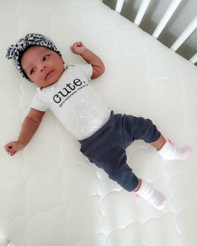 baby laying in crib wearing headband and shirt with word cute on it