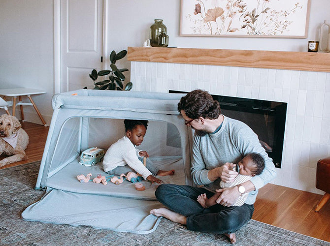 Dad watching toddler play in a potable crib while holding newborn