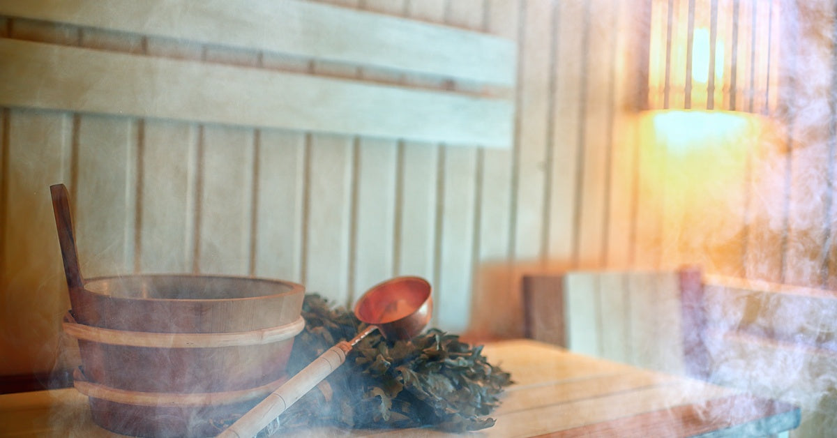 The inside of a small sauna filled with steam. In the foreground, a bucket and ladle sit on a bench.