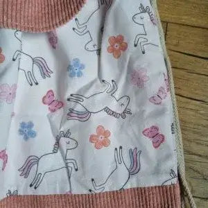 Zoom sur le tissu imprimé d’un sac à dos pochon fait main, orné de licornes, fleurs et papillons dans les tons roses, violets et orangés sur fond rose pâle. Partie inférieure en velours côtelé rose et cordon écru visible sur le côté. Photo prise à plat sur un sol en bois clair.