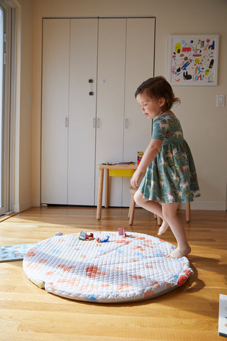 Little girl in green dress, dancing on top of a portable cushioned baby mat with the sun shining in.