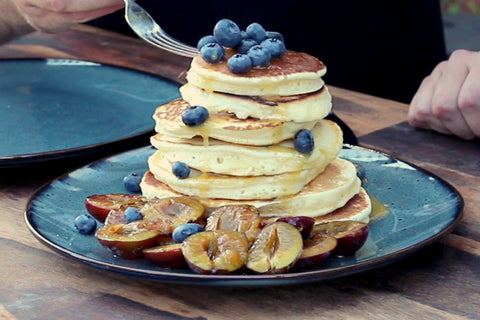 Übereinander geschichtete Pancakes auf einem Teller, die mit Zwetschgen und anderem Obst angerichtet sind.