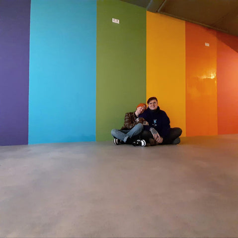 Valentijn and Thomas sitting on the floor cuddling each other in front of a giant painted rainbow wall inside the bookstore.