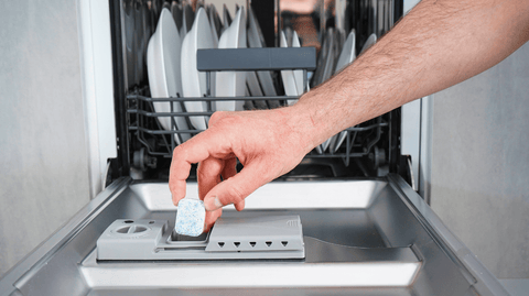 Hand van een persoon plaatst een vaatwastablet in de dispenser van een open vaatwasser gevuld met witte borden.