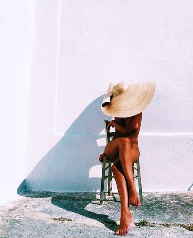 Rear view of a relaxed woman sitting on a wooden chair in Sahra.Nko swimwear, wearing a large straw hat against a white wall, symbolizing tan maintenance.