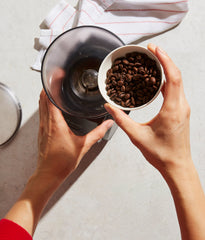 Two hands holding a cup of coffee beans over a coffee grinder.