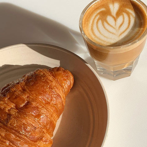 Croissant on a plate next to a flat white with a flower design in the foam.