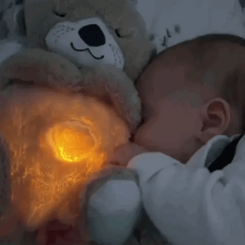 The Snuggle Otter lies next to a baby in the bedroom. The baby cuddles with our Snuggle Otter cuddly toy and the breathing function and lamp are on.