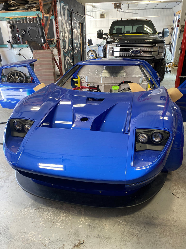 A striking blue sports car in the midst of a B-Quiet Sound Deadening upgrade. The vehicle's hood is open, exposing the engine bay prepared for soundproofing, while the interior is ready for the installation of B-Quiet's noise-reducing materials. The car is set against a backdrop of a busy garage, indicating an active sound deadening project. This setting exemplifies the practical application of B-Quiet's sound deadening solutions, designed for car enthusiasts looking to enhance their vehicle's acoustic comfort.