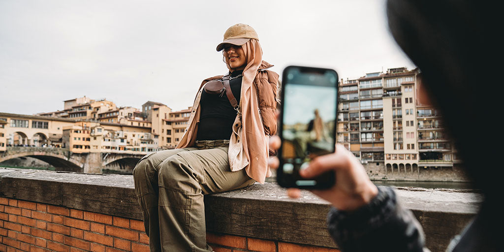 Young woman sits on the ledge of a balcony, posing for a photo. Photo is being taken by a friend holding an iPhone.