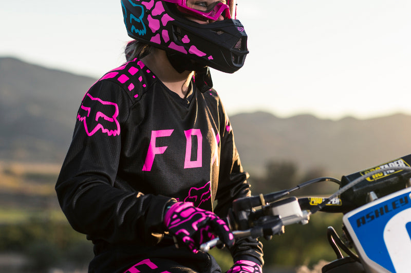 motocyklistka stoi na torze do motocrossu w bluzie Fox 180, ręce oparte o manetki motocykla