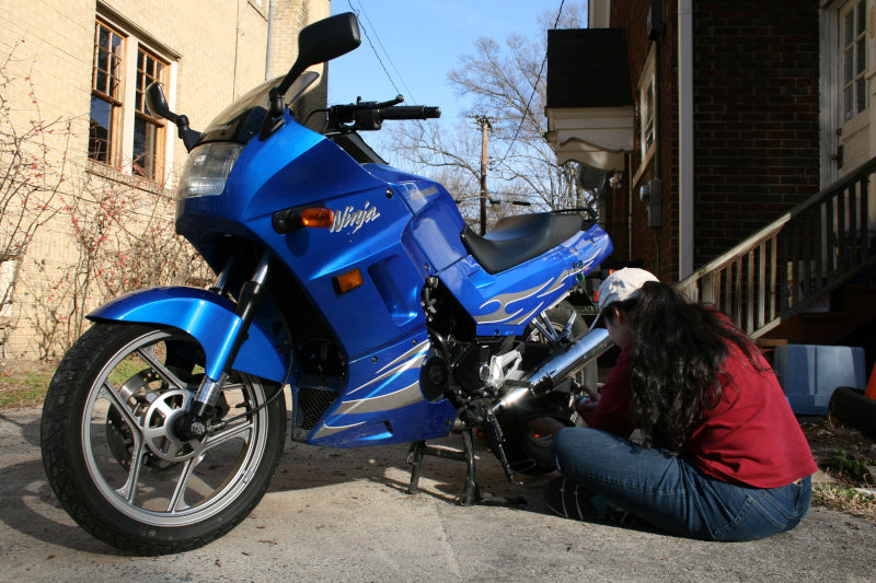 Niebieski motocykl przy budynkach obok kobieta smarująca łańcuch motocykla