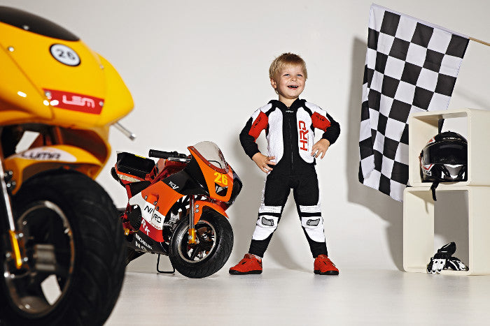 dziecko ubrane w strój motocyklisty stoi przy dwóch małych motorach 