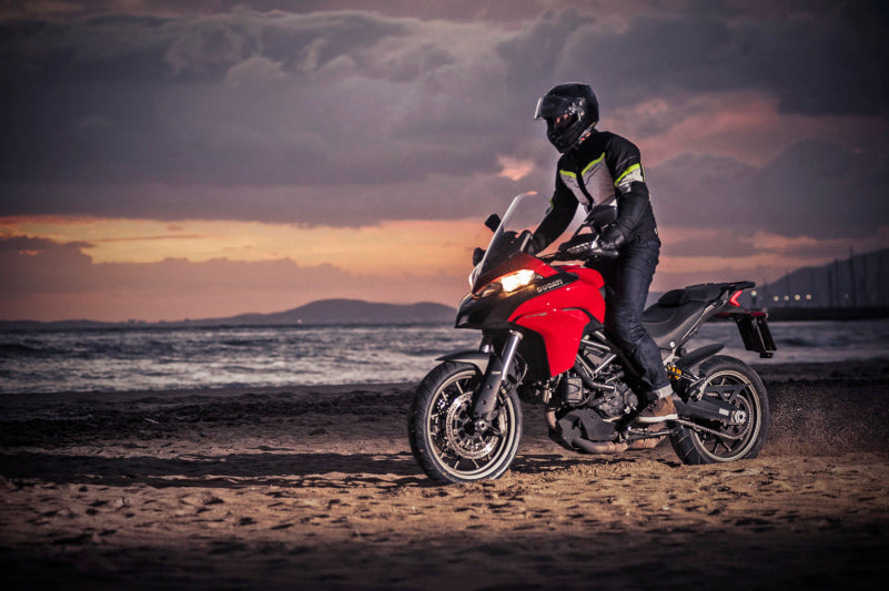 Motocyklista na czerwonym motocyklu ubrany w jeansy i kurtkę marki Rebelhorn na plaży o zachodzie słońca